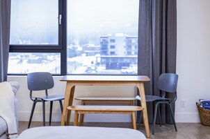 A room with a bed, two chairs, a small wooden table, and a view of buildings through a window.