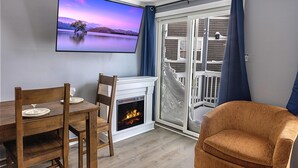 Un salon avec une cheminée, une table à manger avec des chaises, un téléviseur fixé au mur et une vue à travers une porte vitrée.
