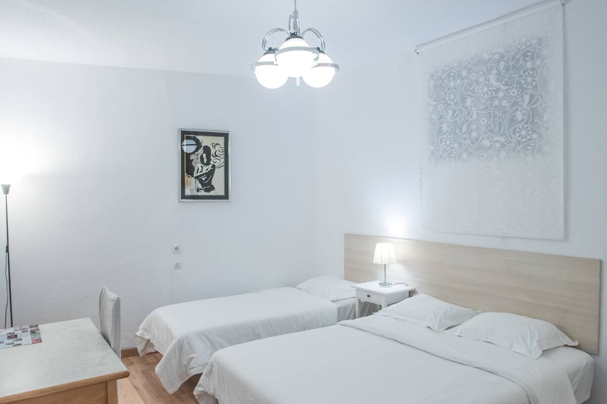 Une chambre d’hôtel avec deux lits simples, un bureau en bois, une chaise, une table de chevet avec une lampe, un luminaire au plafond et une œuvre d’art murale.