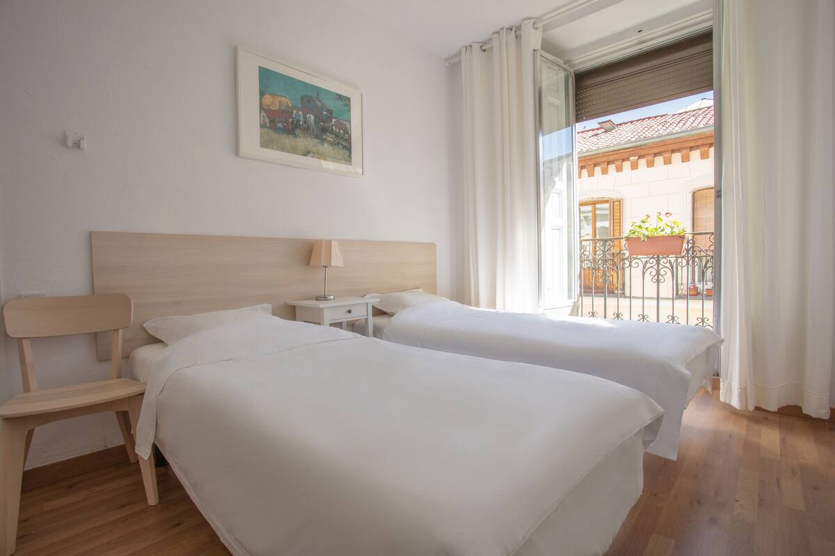 Une chambre d’hôtel avec deux lits, une tête de lit en bois, une table de chevet, une chaise et une fenêtre avec des rideaux.