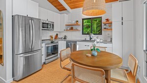 A modern kitchen with white cabinetry, stainless steel appliances, a wooden dining table, wicker chairs, and a hanging light fixture. - Island Chic - Zen Retreat - A/C in 2 Bedrooms (Princeville)
