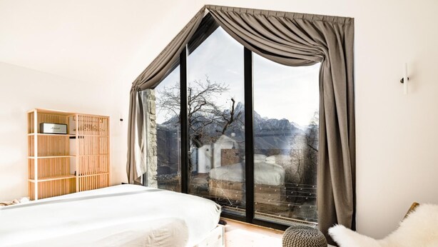 A bedroom with a large window, a bed, a wooden wardrobe, and a white chair. - Chalet with sauna H 311 (GORDONA)