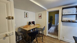 Eine kompakte Küche mit einem Holztisch, Barhocker und einer Kaffeemaschine. Eine Tür führt in einen Essbereich mit einem gelben Stuhl und einer Pflanze.