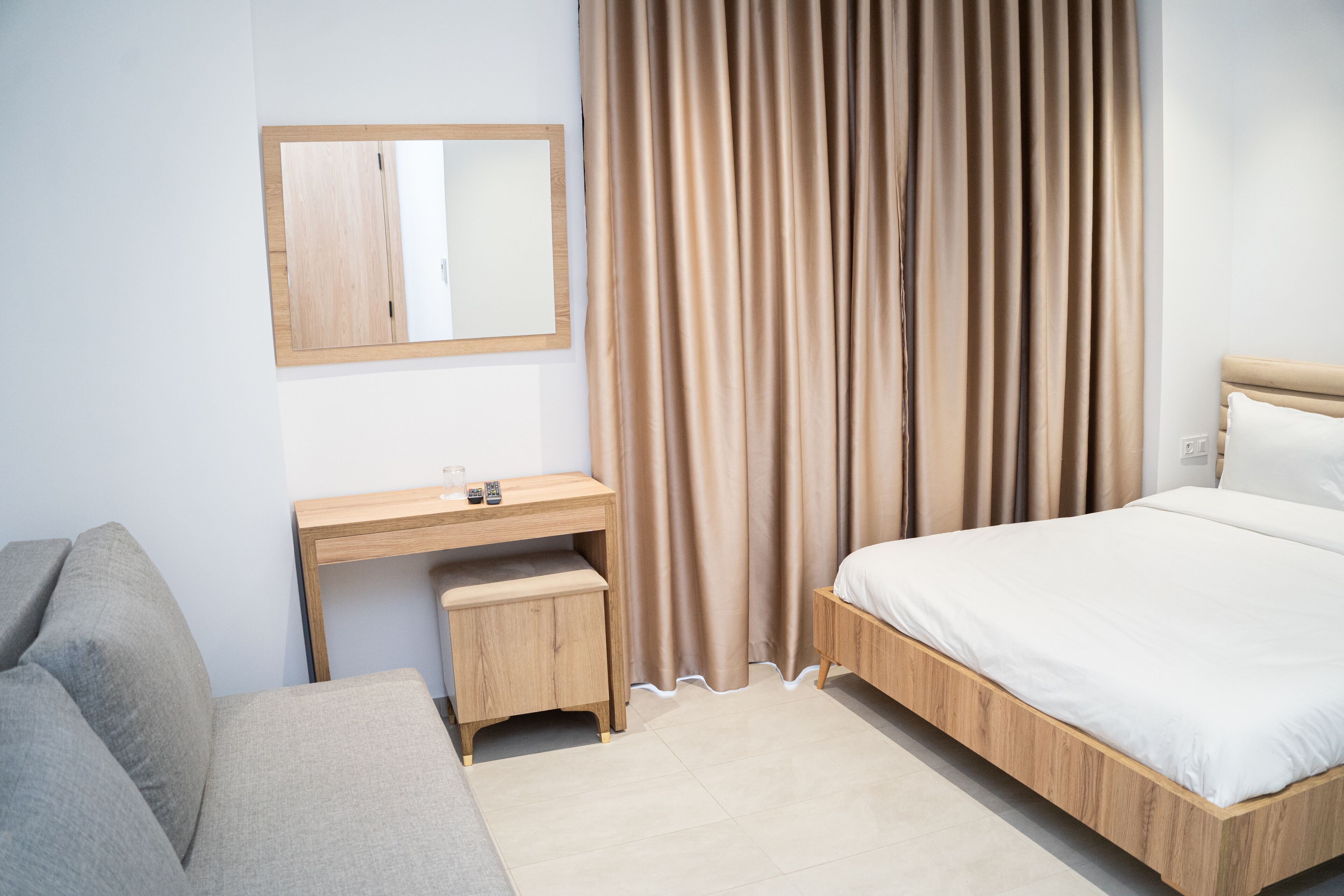 A modern hotel room with a wooden desk, a small stool, a bed with a wooden frame, and a large window with curtains.