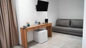 Una habitación moderna con un escritorio de madera, un refrigerador blanco pequeño, un televisor montado, un sofá gris y una cortina.