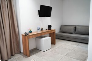 Una habitación moderna con un escritorio de madera, un refrigerador blanco pequeño, un televisor montado, un sofá gris y una cortina.