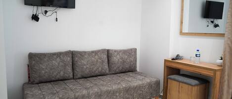 Una habitación con un sofá gris, un escritorio de madera con un espejo, un televisor montado en la pared y un taburete pequeño.