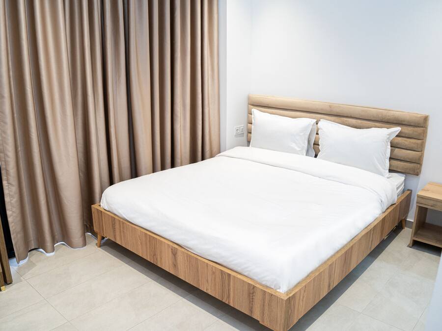 Una cama bien tendida con estructura de madera y ropa blanca, una mesita de noche de madera a su lado, y una cortina ligeramente abierta.