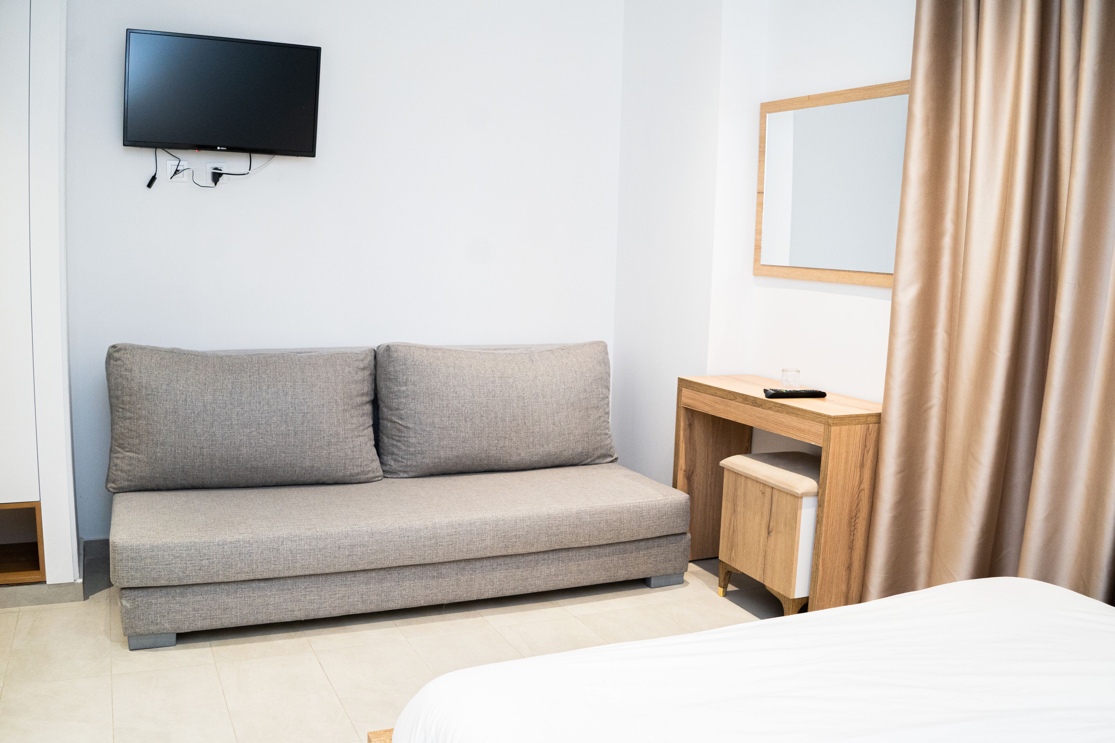 A hotel room with a grey sofa, a wooden desk, a television mounted on the wall, and a bed with a white duvet.
