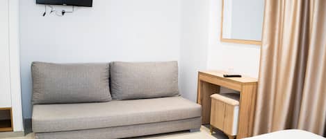 Habitación de hotel con sofá gris, escritorio de madera, televisor montado en la pared y cama con edredón blanco.