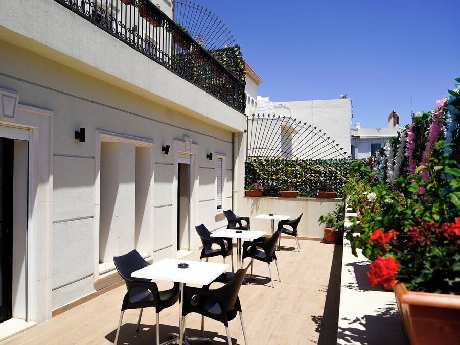 An outdoor seating area with tables and chairs, a white building with balconies, and potted plants.