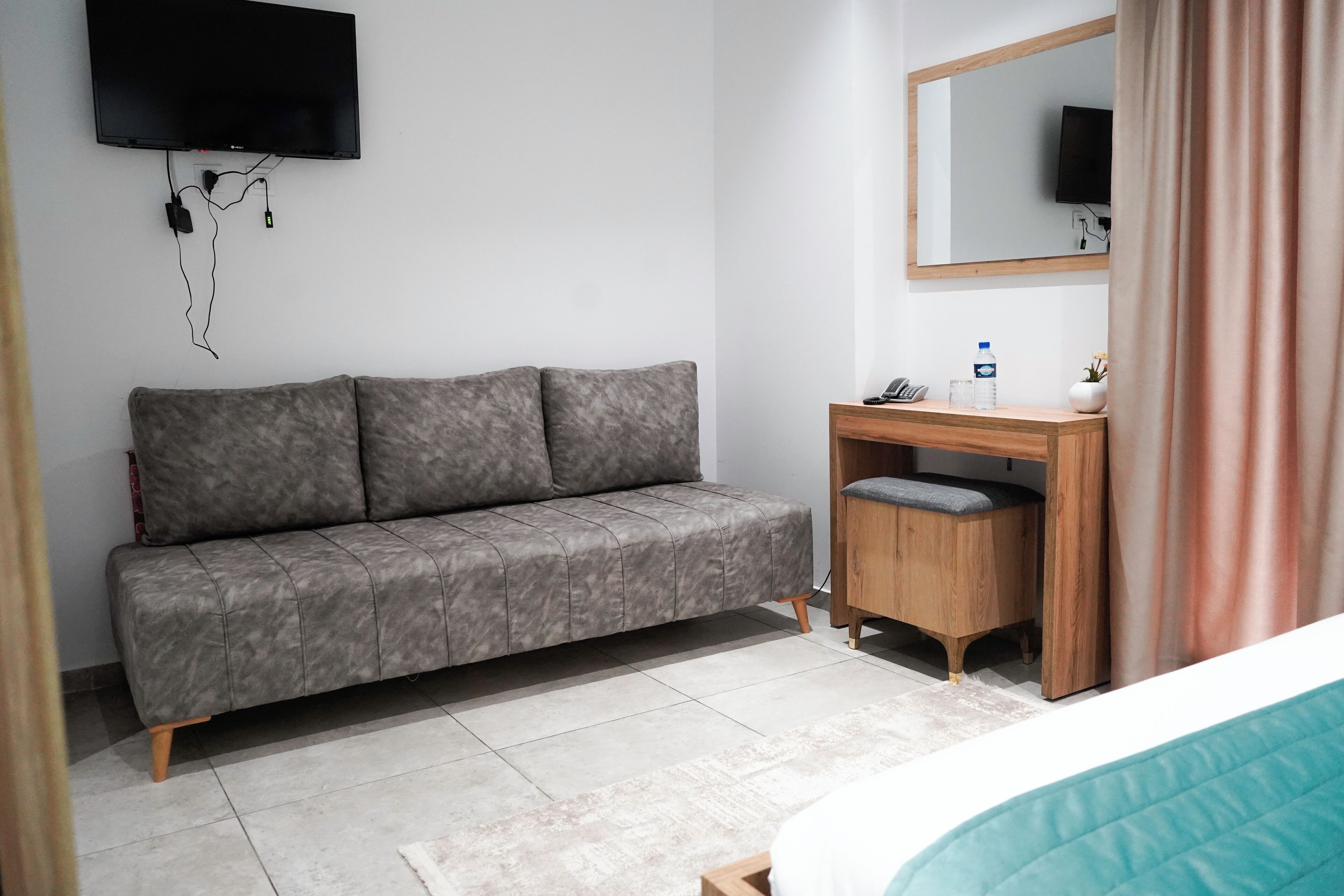 A modern hotel room with a grey sofa, a wooden desk, a mirror, and a television mounted on the wall.
