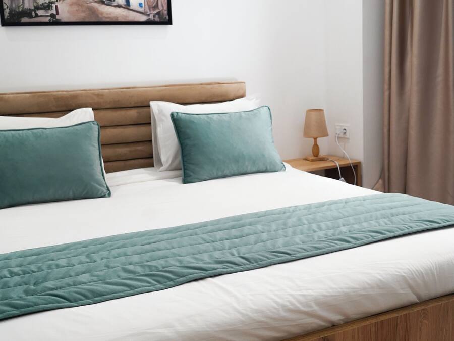 Un lit bien fait, avec une tête de lit en bois, un plaid turquoise et deux coussins turquoise. Un tableau représentant une scène de rue est accroché au mur au-dessus du lit.