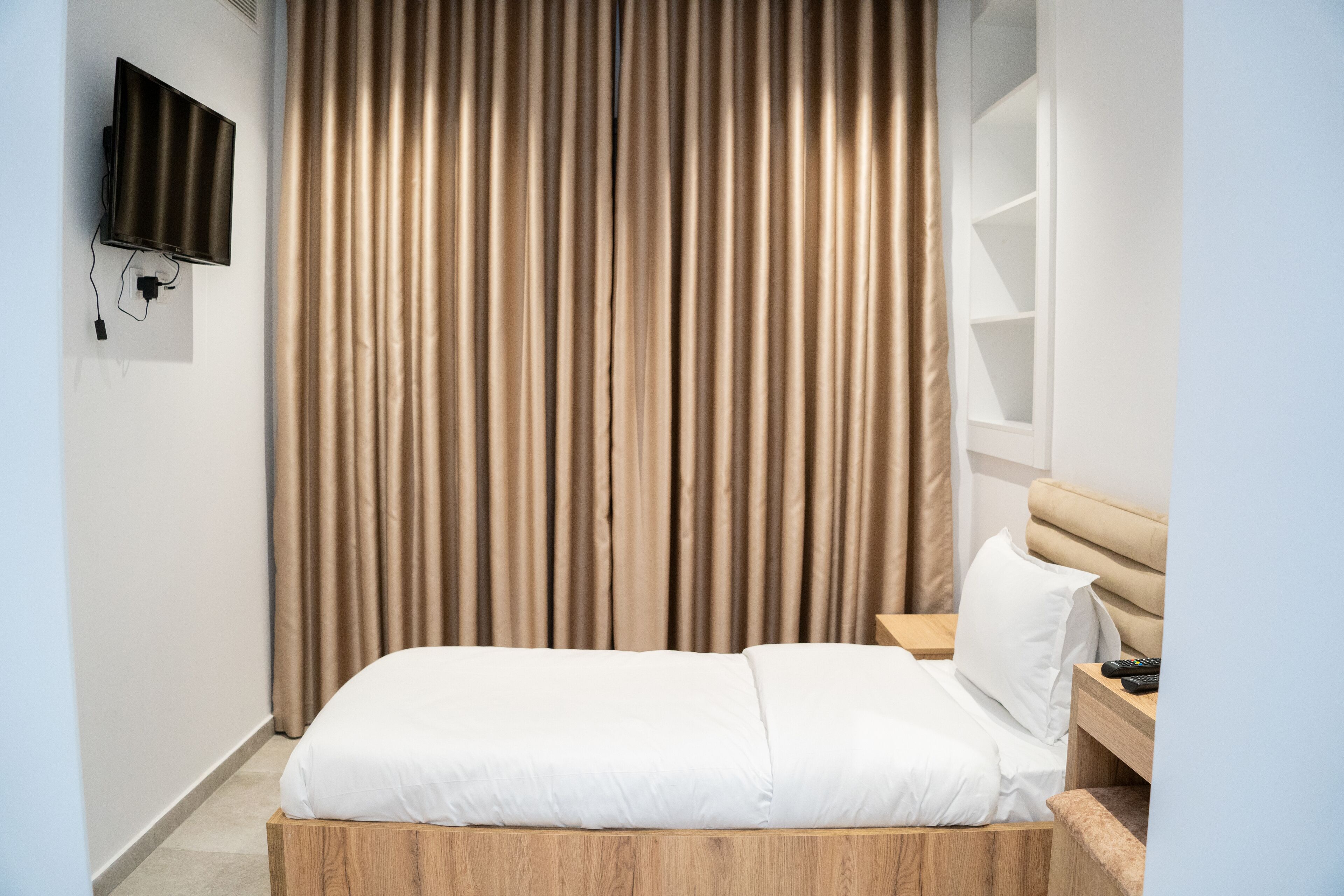 Une chambre d’hôtel avec un lit, une tête de lit en bois, un téléviseur fixé au mur et un rideau.
