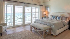 Ein Schlafzimmer mit einem großen Bett, einer Bank, einem Nachttisch und Blick auf den Ozean durch eine Schiebetür.