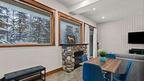 A cozy living room with a stone fireplace, a wooden dining table, blue chairs, and a grey sofa. - Lrg 1 Bedroom Suite -Bike Storage-Private Hot Tub! (Sun Peaks)