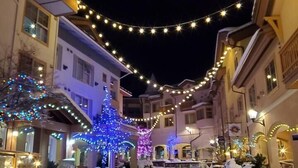 A snow-covered street with buildings on either side, decorated with string lights and illuminated trees. - Lrg 1 Bedroom Suite -Bike Storage-Private Hot Tub! (Sun Peaks)
