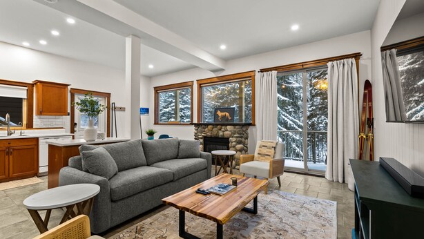 A modern living room with a fireplace, a sofa, a coffee table, and a view of snow-covered trees through large windows. - Lrg 1 Bedroom Suite -Bike Storage-Private Hot Tub! (Sun Peaks)