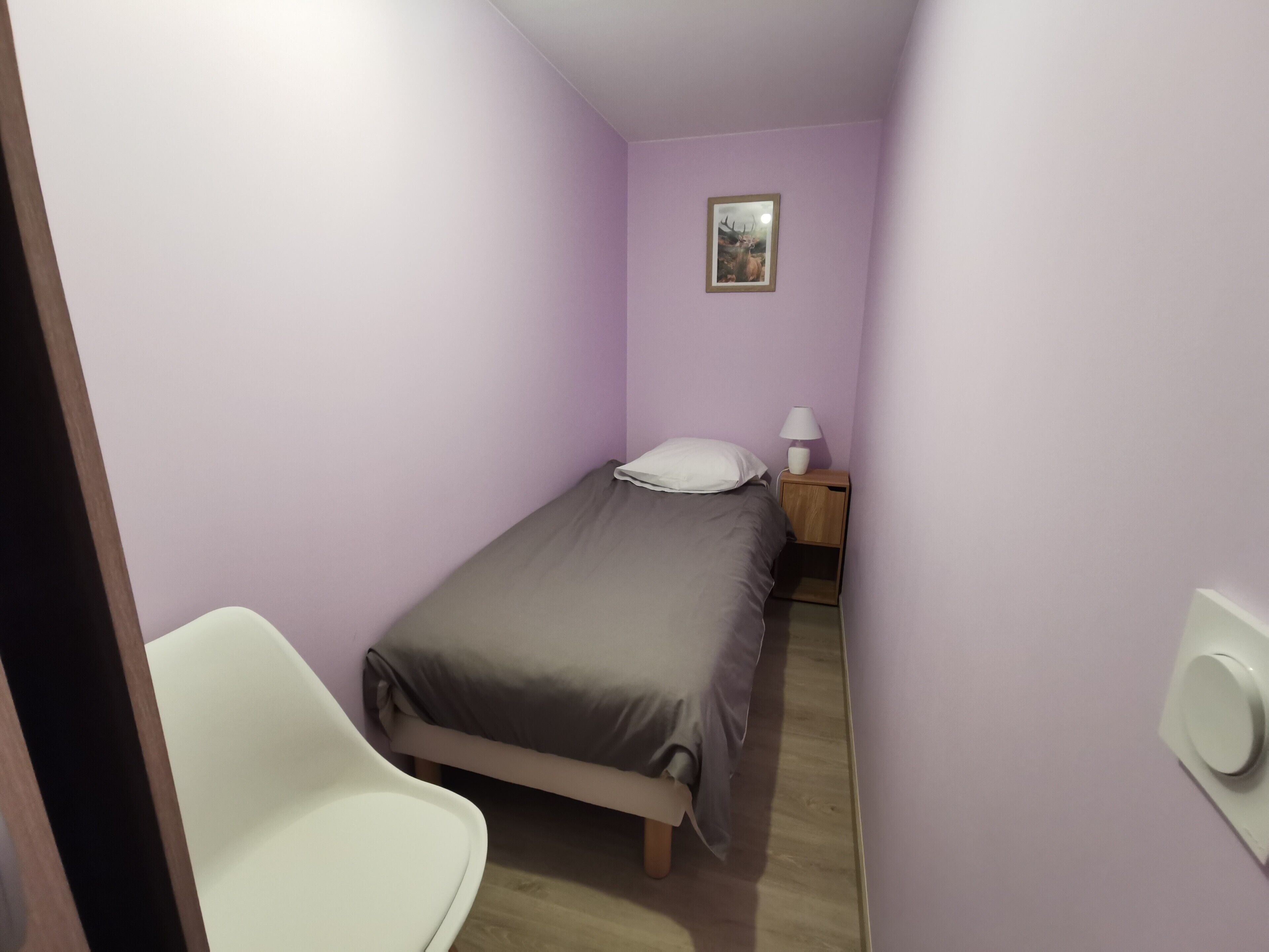 A small, narrow room with a single bed, a nightstand with a lamp, and a white chair.