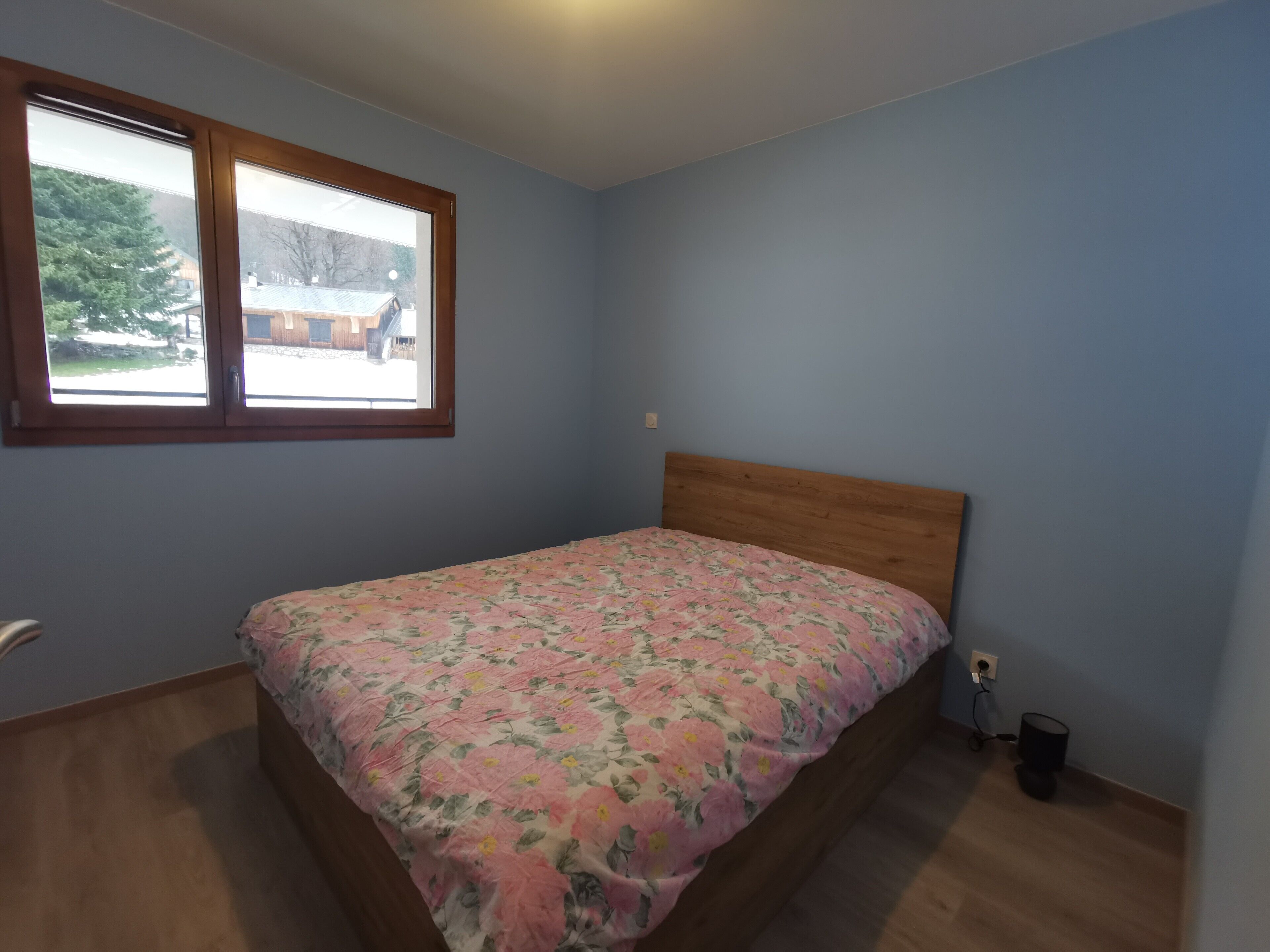 Ein Schlafzimmer mit einem Holz-Kopfteil, einer blumigen Tagesdecke, einem Fenster mit Blick auf ein Haus und Bäume sowie einem kleinen Nachttisch mit Lampe.