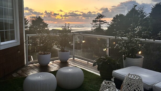 En terrasse med udendørs møbler, plantekasser og udsigt til havet i solnedgang.