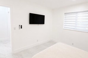 A room with a flat-screen TV mounted on the wall, a window with blinds, and a white door.