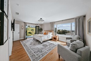 Un dormitorio con una cama grande, un sofá gris, suelo de madera y vistas al exterior.
