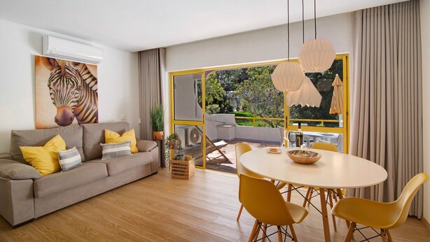A modern living room with a sofa, yellow cushions, a dining table with yellow chairs, and a large window with curtains.
