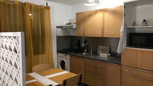 A compact kitchen with wooden cabinets, a washing machine, a microwave, and a dining area with a table and chairs. - Beau Appartement Calme et Ensoleillé Parisdisney (Noisy-le-Grand)