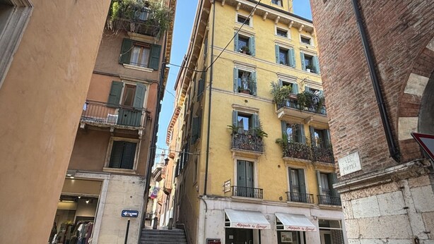 A narrow street with multi-story buildings, balconies with plants, and a visible storefront. - Lux Lodge near Piazza Erbe (Verona)