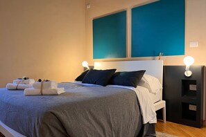A neatly made bed with a grey blanket, two pillows, and a set of towels on the side. There are two wall-mounted light fixtures on either side of the bed. - Lux Lodge near Piazza Erbe (Verona)