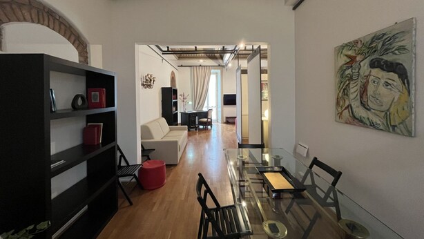 Een moderne woonkamer met een glazen eettafel, zwarte stoelen, een rode poef en een zwarte boekenkast.