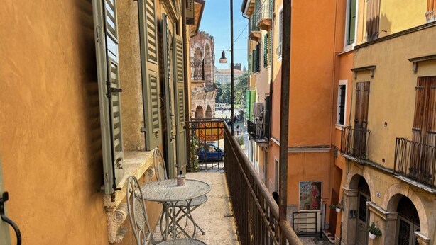 A narrow alleyway with a balcony featuring two metal tables and chairs, a wrought iron railing, and a view of a historic building with arches in the background. - LOFT 17.2 - Verona Journeys (Verona)