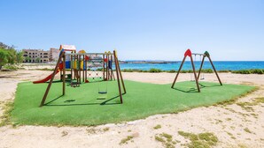 Een speeltuin met schommels en een glijbaan, gelegen aan een zandstrand met een heldere lucht en een gebouw op de achtergrond.