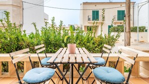 Een terras buiten met een houten tafel en stoelen, omringd door groen en gebouwen op de achtergrond.