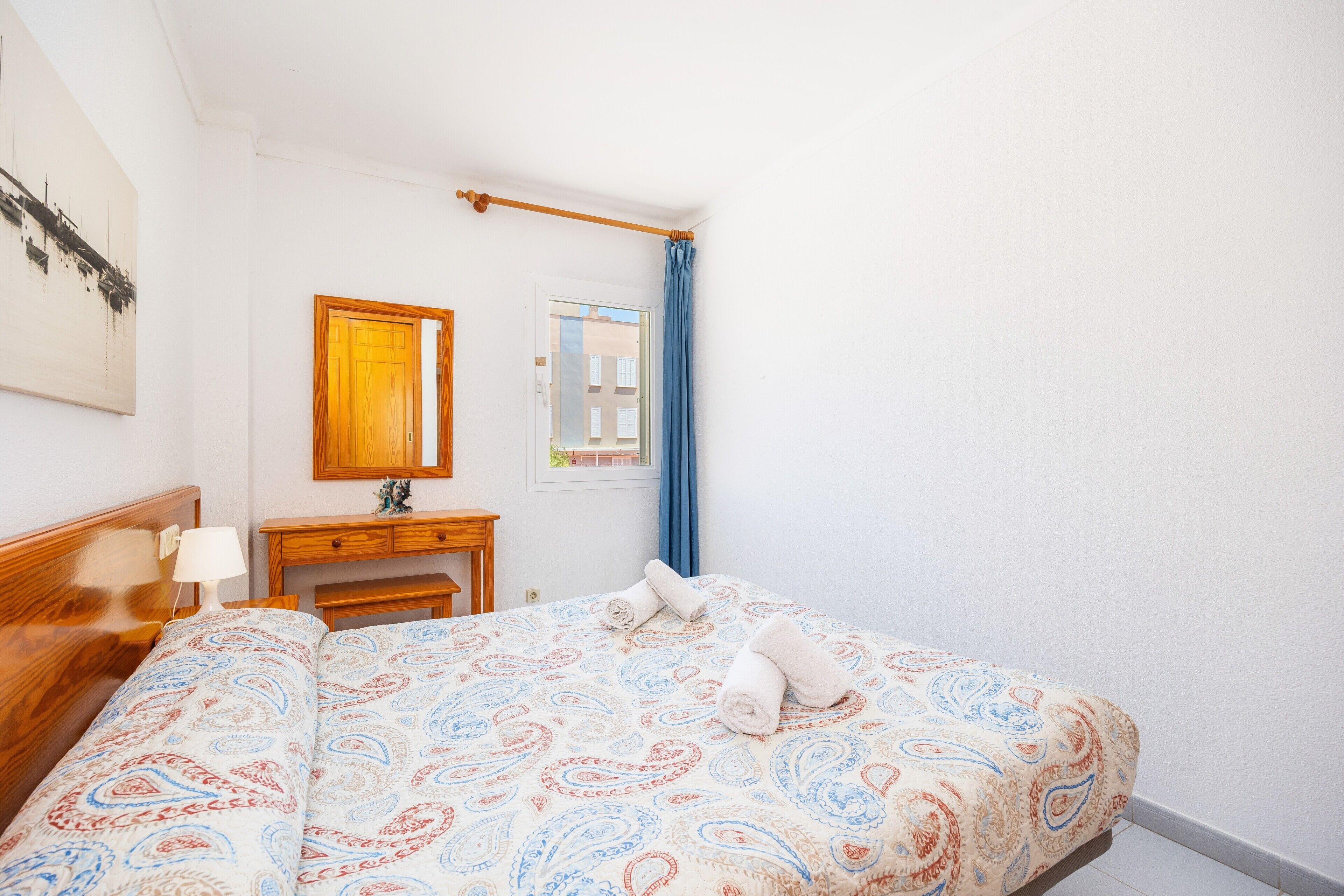 A hotel room with a bed, a wooden headboard, a nightstand, a desk, a mirror, a window with blue curtains, and a wall-mounted artwork.