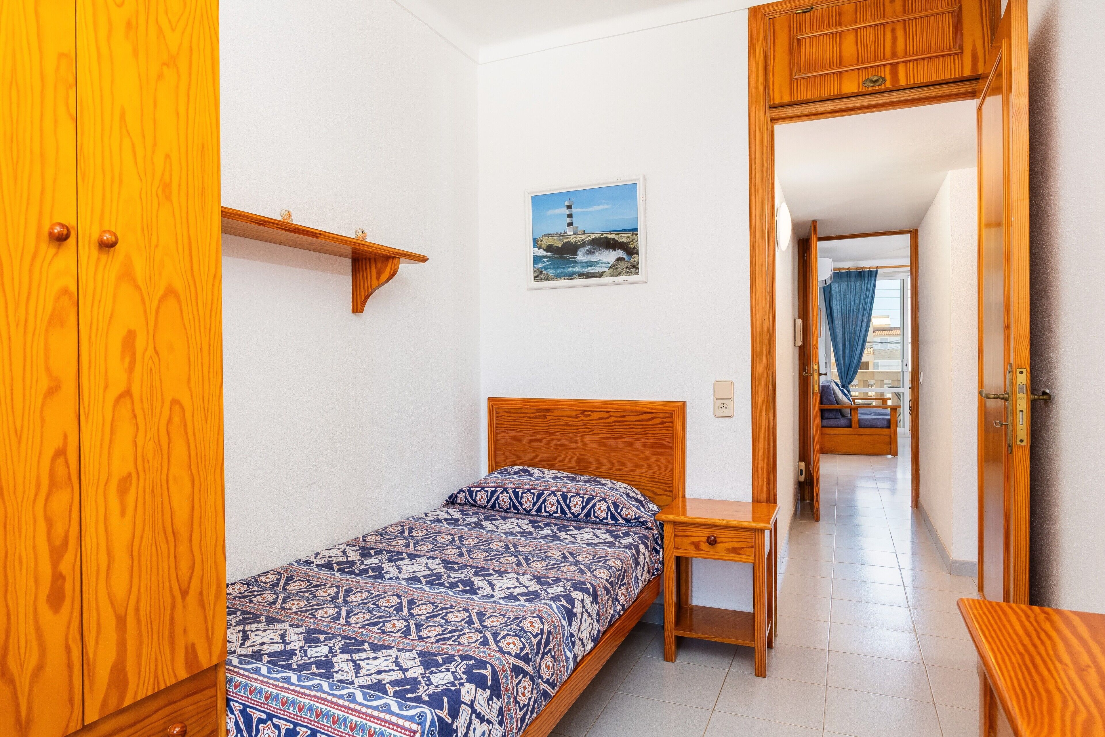 Ein Schlafzimmer mit einem Bett, einem Nachttisch aus Holz, einem Kleiderschrank, einem Bild an der Wand und einem Flur, der in ein weiteres Zimmer führt.