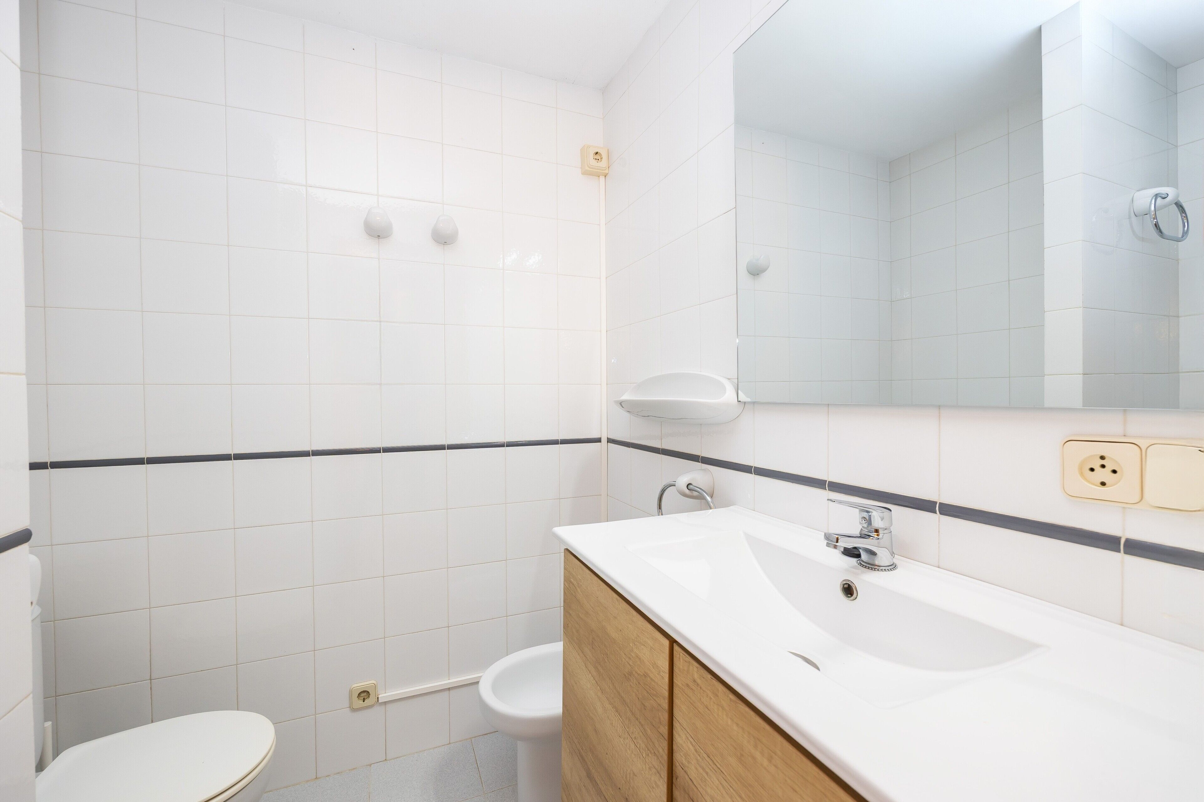 A bathroom with white tiles, a toilet, a sink with a mirror, and a bidet.