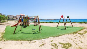 A playground with swings and slides on a sandy area with a clear sky and a building in the background. - Apartment 'Can Vidal Cabrera' with Private Terrace, Close to the Beach (Tourist Taxes Included) (Ses Salines)