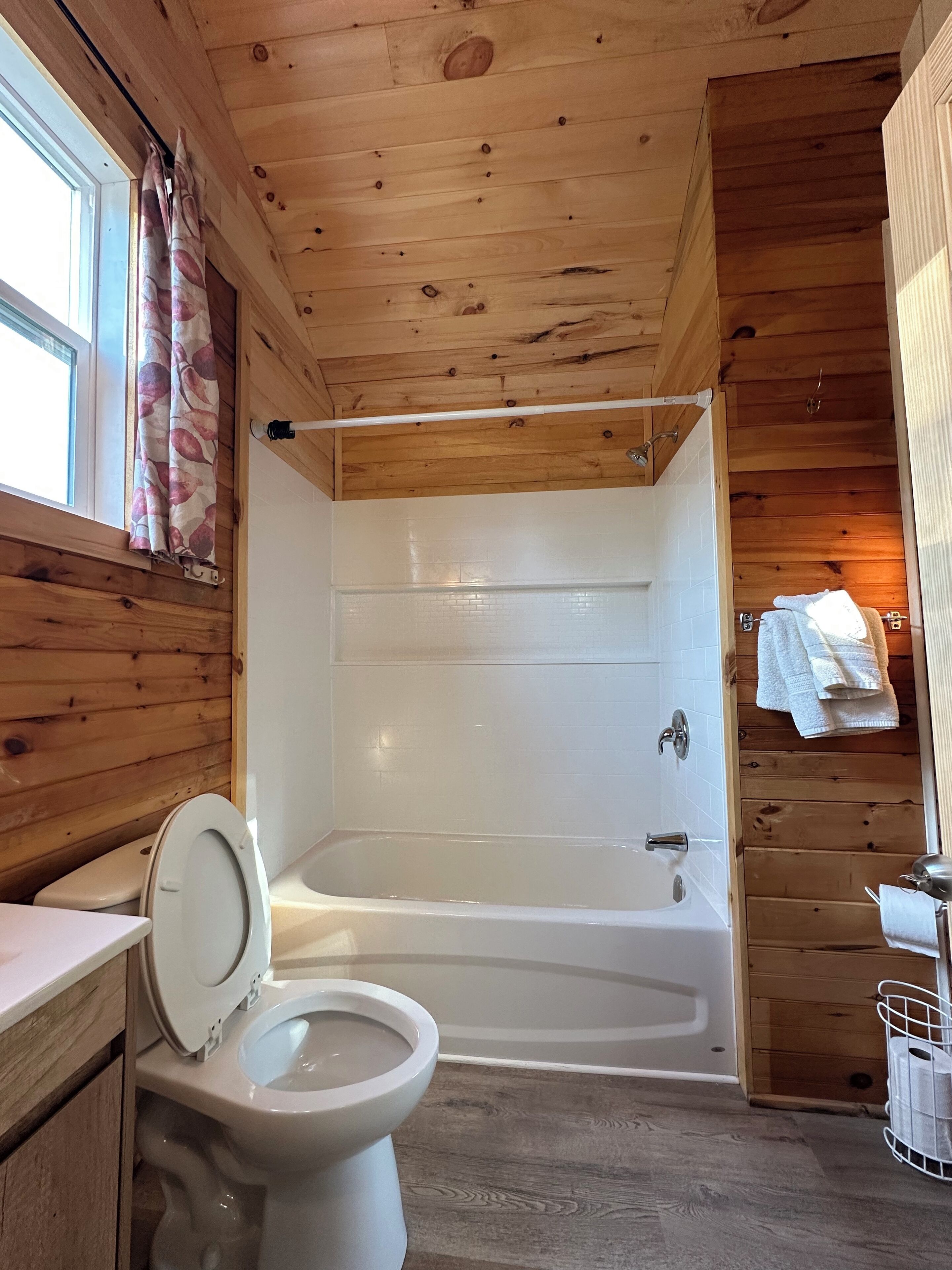Un baño de cabaña de madera con inodoro, bañera y una ventana con cortinas.