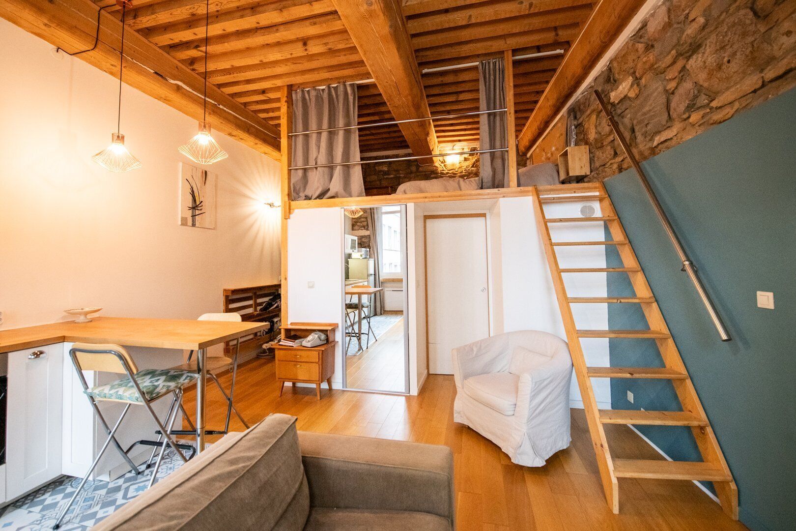 Un accogliente loft con soffitto in legno, una zona pranzo con tavolo e sedie, una scala per un livello superiore e un piccolo angolo relax con una poltrona e un divano.