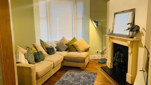 A living room with a beige sectional sofa, a fireplace, and a large window with blinds. - Beautiful 3-bed House in Preston (Preston)