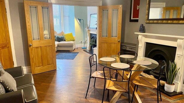 A dining area with a round glass table and wooden chairs, a fireplace, and a sofa in a living room with hardwood flooring. - Beautiful 3-bed House in Preston (Preston)