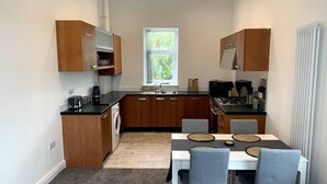 Kompakti keittiö, jossa on puiset kaapit, sisäänrakennettu uuni sekä ruokailutila, jossa on pöytä ja tuolit.