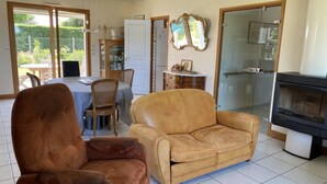 Ein Wohnzimmer mit einer Couch, einem Sessel, einem Esstisch mit Stühlen, einer Küchenzeile mit Herd und einer Glastür zum Garten.