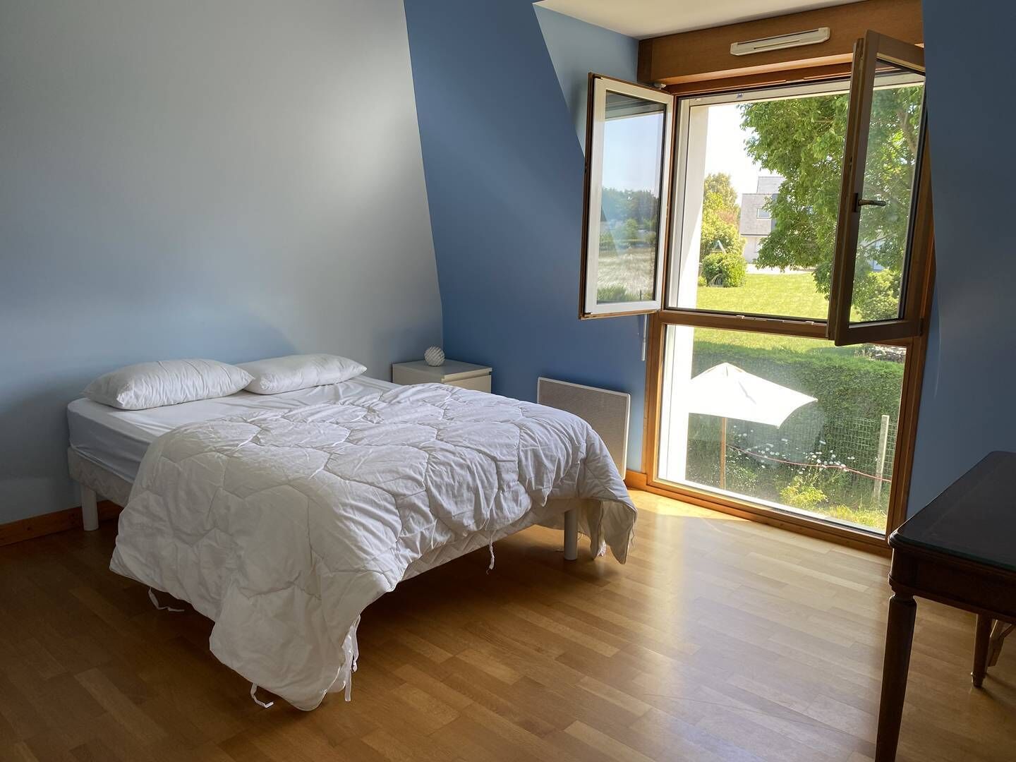 A bedroom with a bed, a nightstand, a window with an open blind, and a view of greenery outside.