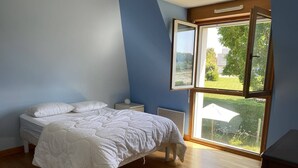 Ein Schlafzimmer mit einem Bett, einem Nachttisch, einem Fenster mit geöffneter Jalousie und Blick auf Grünfläche.