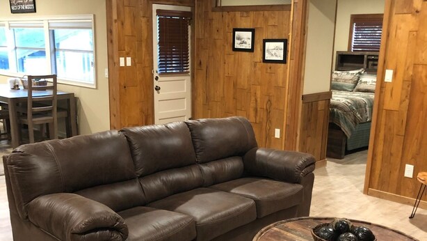 Un salon avec un canapé en cuir marron, une table basse ronde en bois agrémentée d’objets décoratifs, des murs recouverts de panneaux en bois, un coin repas avec une table et des chaises, et une chambre à coucher avec un lit et des stores aux fenêtres.