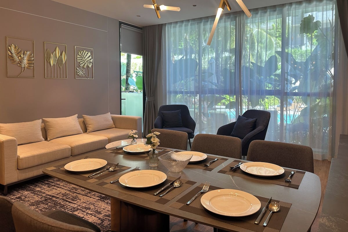 A modern dining area with a long table set for four, a sofa, and a view of a pool through large windows.