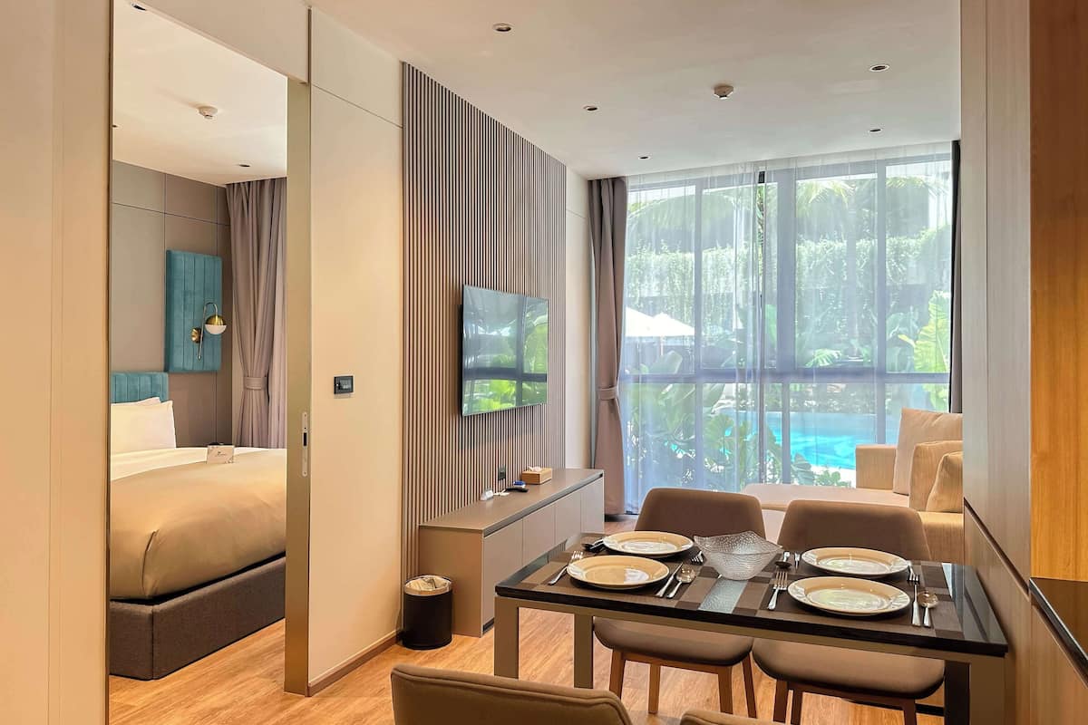 A modern hotel room with a bed, a TV mounted on the wall, a dining table set with plates and cutlery, and a view of a swimming pool through large windows.